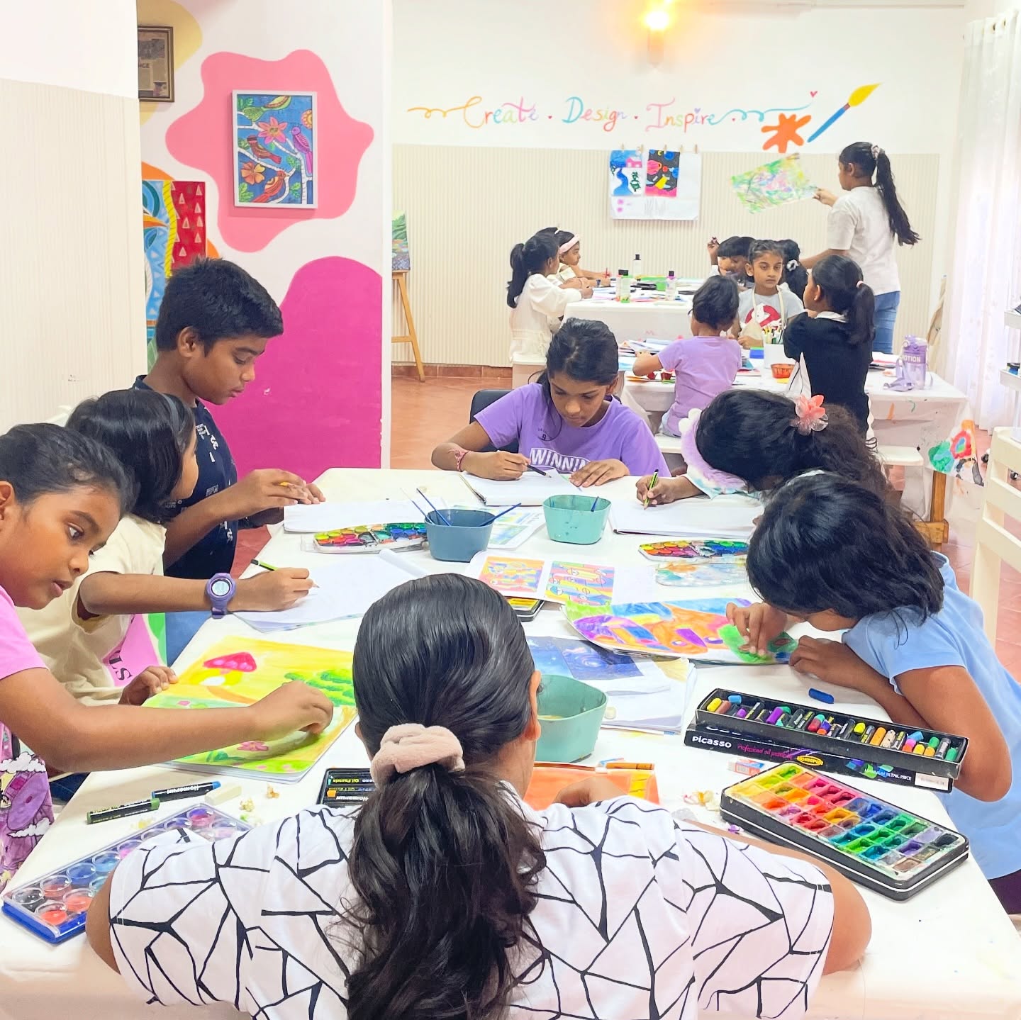 Kids painting and drawing at tables in the Art Club studio, with finished artwork pinned to the wall behind them