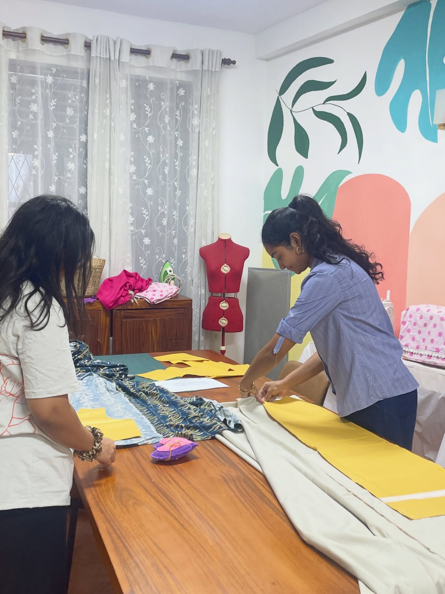 Two women pattern-cutting and pinning fabric at a sewing table beside a dress form in the studio