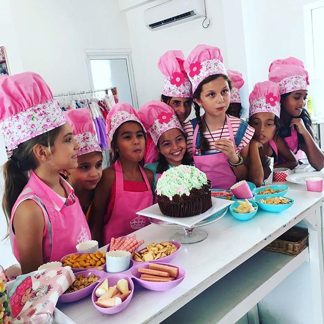 A group of kids in pink chef hats and aprons gathered around a decorated cake at a baking party