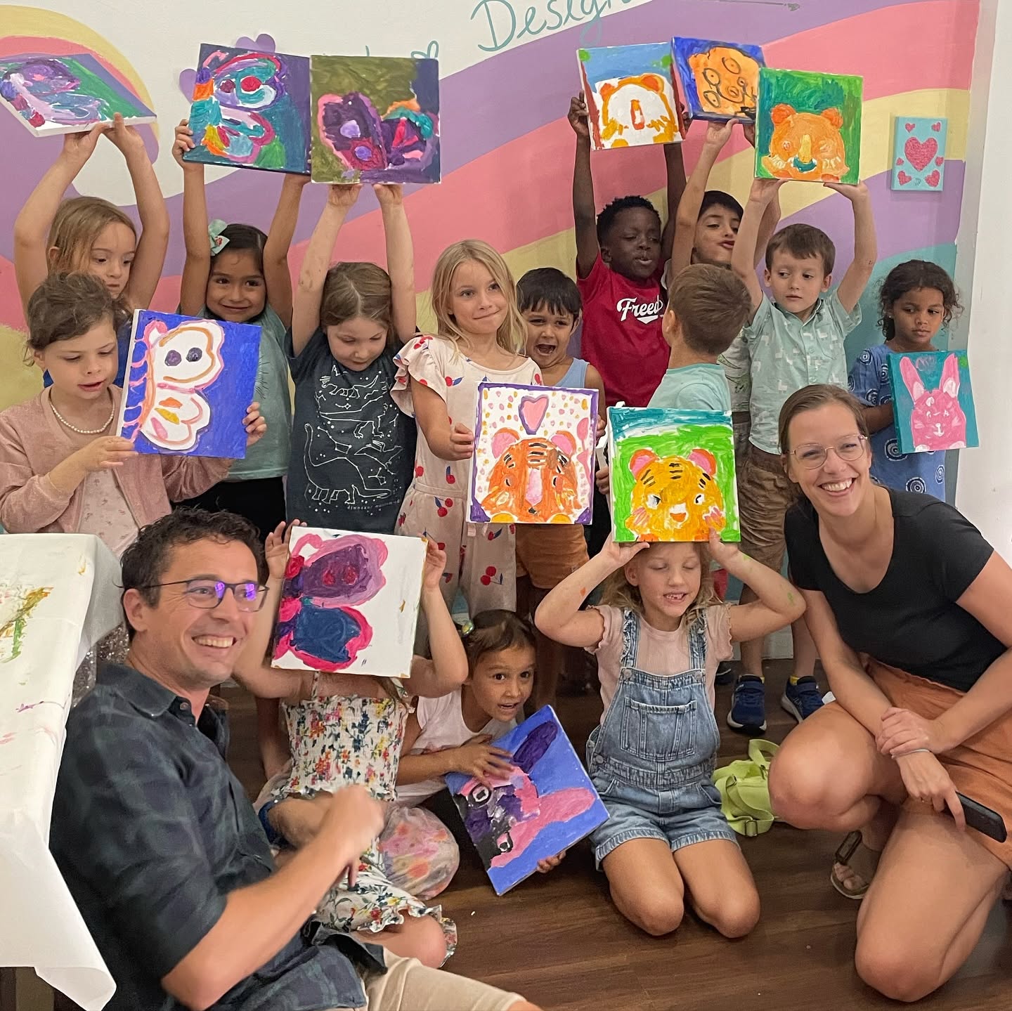 A large group of kids and two teachers holding up their painted animal canvases in the art studio